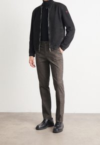 Giacca di denim nera con colletto alto, dolcevita grigia, pantaloni marroni con texture, e scarpe in pelle nere su uno sfondo neutro.