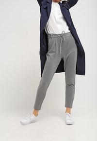 Grijze joggerbroek met een elastische tailleband en trekkoord, gecombineerd met een witte top en een lange marineblauwe jas. Witte sneakers maken de look compleet.