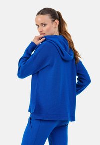 Sweat à capuche bleu en tissu maille doux, avec capuche à cordon, coupe décontractée et poignets côtelés. Porté avec un bas assorti.