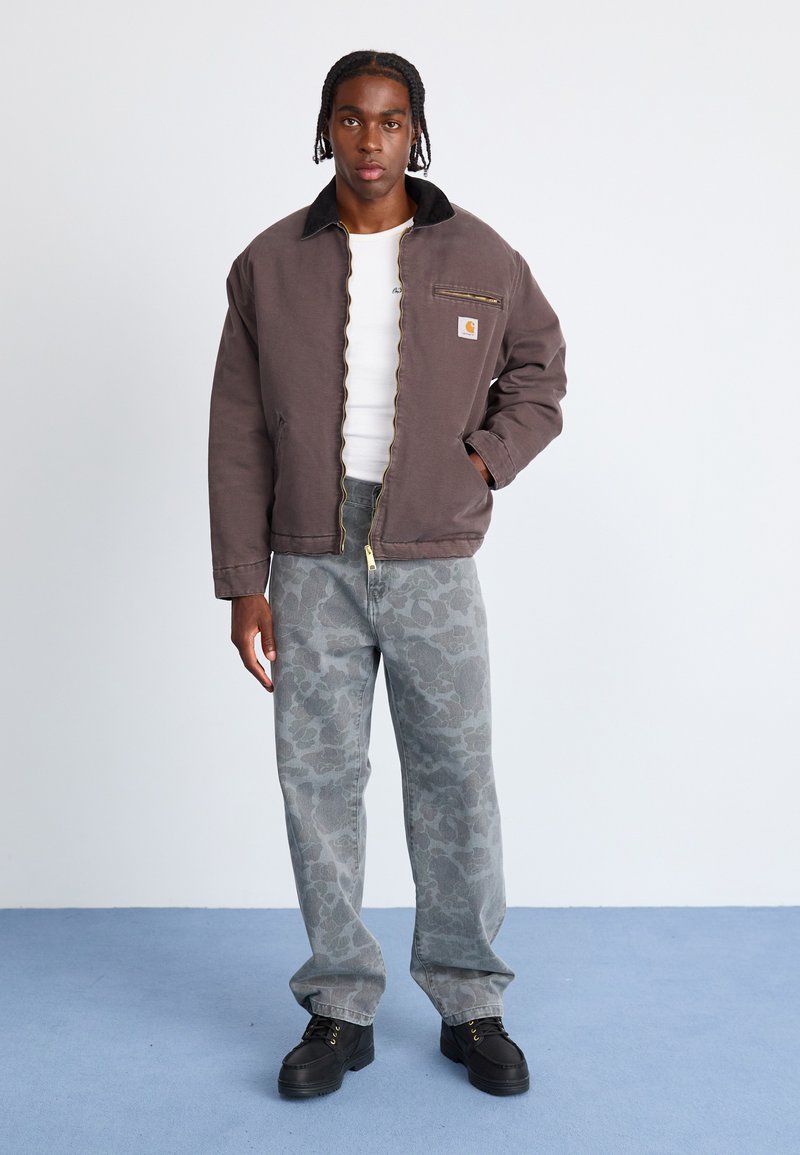 Bruine geïsoleerde jas met zwarte kraag, witte shirt eronder, grijs gemarmerde broek en zwarte laarzen, staande tegen een lichte achtergrond.