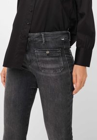 Jeans en denim noir avec une texture lisse, dotés d'un design à cinq poches et de coutures blanches subtiles. Fermeture par bouton et zip à la taille.