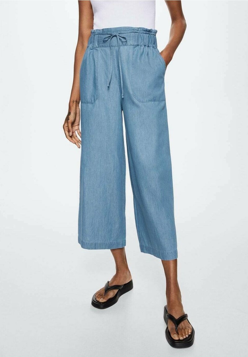 Femme portant un pantalon cropped large taille haute de couleur bleu clair avec cordon de serrage et poches avant, associé à des tongs à plateforme noires.