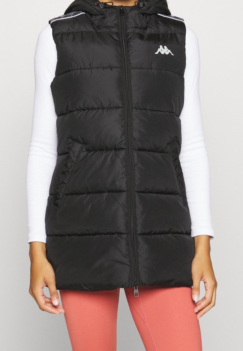 Veste noire matelassée sans manches avec capuche, fermeture éclair devant et poches, portée sur un haut blanc à manches longues et un pantalon corail.