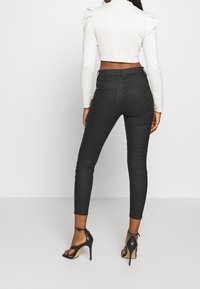 Jeans negros ajustados con bolsillos traseros y costuras texturizadas, combinados con una blusa corta de color crema y tacones negros destalonados.