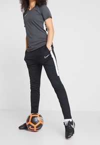 Person, die schwarze und graue Nike-Sportbekleidung trägt, steht mit den Händen in den Taschen, ein Fuß ruht auf einem schwarz-orangefarbenen Fußball.