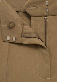 Beige tyg med struktur, försett med dragkedja, knapp och två metalldetaljer. Har dekorativ stickning och en ficköppning på sidan.