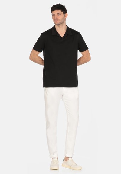Polo shirt nera con colletto classico, maniche corte e design slim, abbinata a pantaloni bianchi e scarpe da ginnastica chiare.