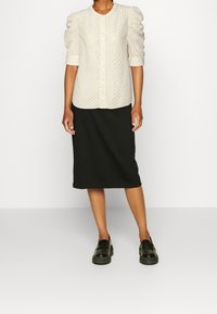 Femme portant un chemisier crème à manches bouffantes avec des boutons, une jupe noire longueur genou et des mocassins noirs, debout devant un fond blanc uni.