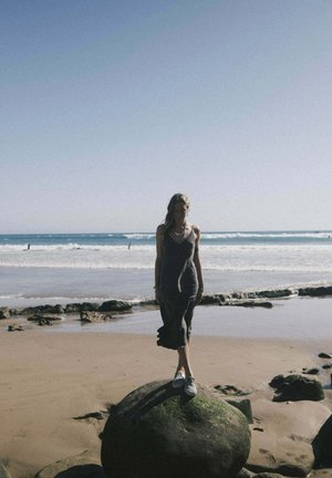 Długa szara sukienka z falbaną u dołu, noszona z kratkowymi butami typu slip-on, stojąca na dużym kamieniu na plaży, z falami w tle.