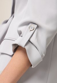 Light gray blouse fabric with a smooth texture. Rolled sleeve featuring a silver button accent for adjustable fit.