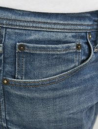 Sinine denim-püksid, millel on kulunud tekstuur, väike tagatasku, messingist nupud ja kontrastsed oranžid õmblused.