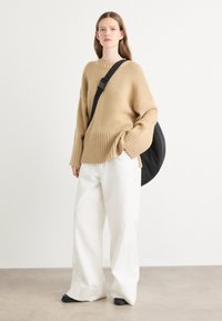 Beige oversized gebreide trui met geribbelde onderkant, gecombineerd met witte wijde broek en een zwarte schoudertas. Glad, zacht textuur.