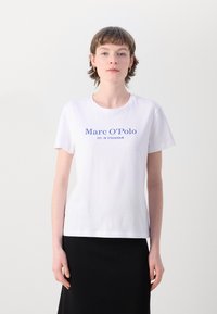 Balta kokvilnas t-krekls ar zilu tekstu "Marc O'Polo EST. STOCKHOLM" priekšā, apaļa kakla izgriezuma un īsām piedurknēm. Redzama melna svārki.