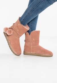Růžové semišové kotníkové boty s umělou kožešinou, strukturální povrch a plochou gumovou podrážkou. Mají detaily se stuhou po straně a otevřený vršek.