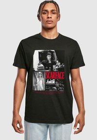 Sort bomulds t-shirt med "Scarface" grafik, der viser sort-hvide filmstill fra filmen og rød tekst i bunden. Klassisk rund hals design.