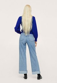 Haut bleu à manches longues avec des épaules bouffantes, associé à un jean large bleu clair. Le jean présente des ourlets effilochés et une coupe décontractée. Chaussures noires.