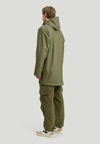 Olivenfarvet vandtæt jakke med hætte, lange ærmer og sidesømme. Parret med olivenfarvede cargo-bukser og beige sneakers.