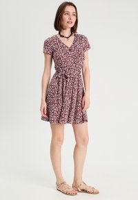 Robe bordeaux à manches courtes avec un motif floral, taille cintrée et jupe évasée. Associée à des sandales beiges à lanières et un collier simple.