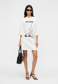 Robe t-shirt blanche surdimensionnée avec un graphisme représentant un canard portant des baskets et une chaîne. Accessoirisée avec des lunettes de soleil noires et un sac à main noir.