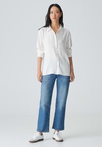 Chemise blanche à manches longues associée à un jean large bleu clair et des baskets blanches, sur un fond neutre.
