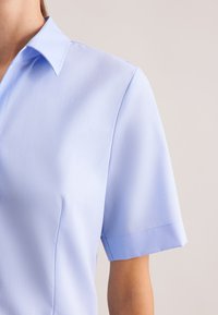 Camicia azzurro chiaro a maniche corte con colletto classico, texture liscia e design su misura con dettagli di cuciture sottili.