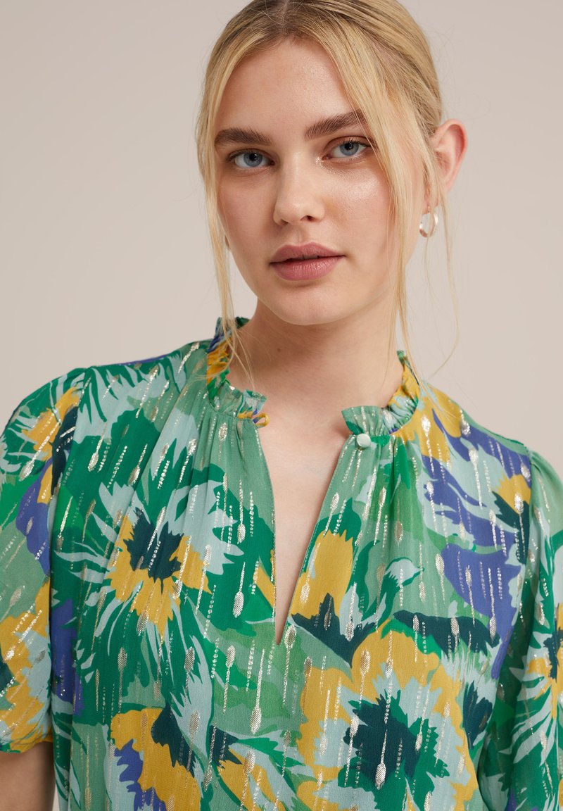 Bluse mit floralem Muster in Grün, Gelb und Blau, aus weichem Stoff, mit Stehkragen und Knopfdetails sowie dezenten metallischen Akzenten.
