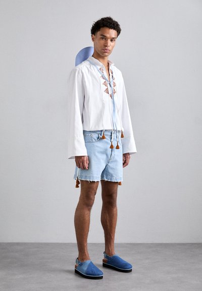 Chemise blanche brodée avec des accents bleus, short en denim clair avec des franges, et chaussures slip-on bleues. Tenue moderne décontractée avec des textures superposées.