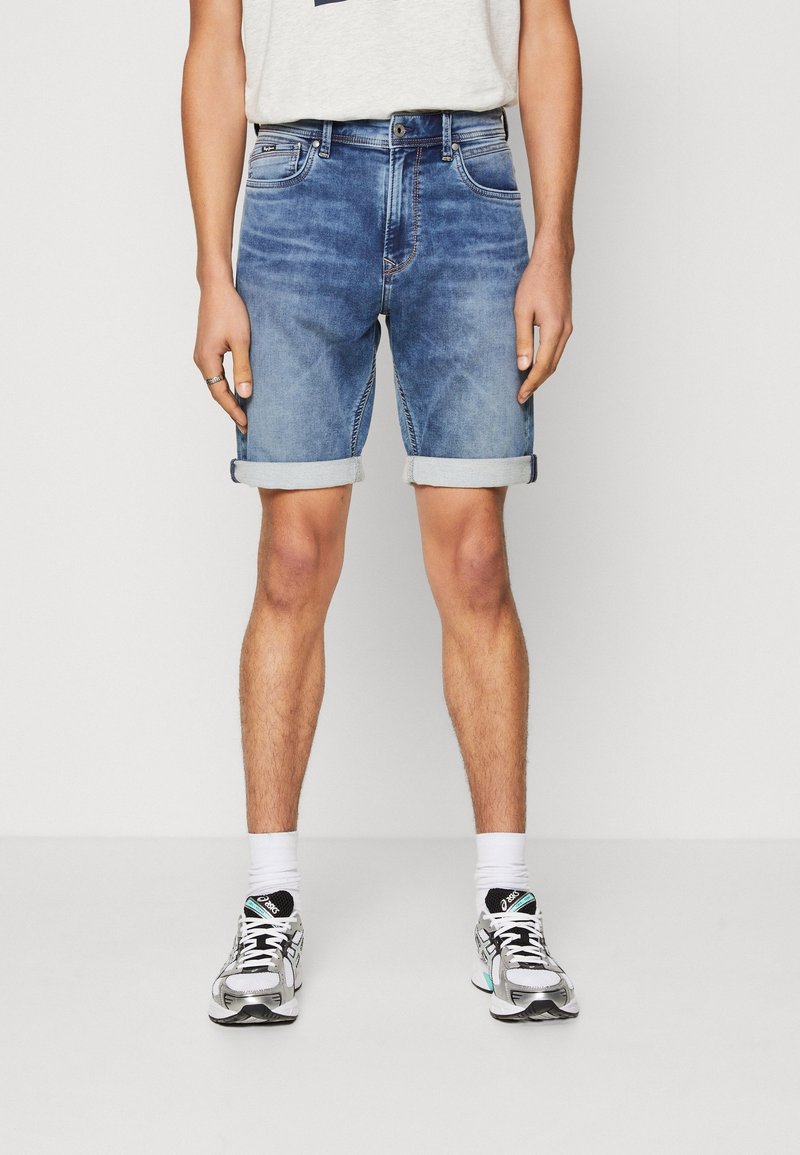 Mies, joka pukee ylös käärityt siniset farkkushortsit, valkoisen T-paidan, valkoiset sukat ja hopeiset juoksukengät, seisoo yksinkertaisella valkoisella taustalla.