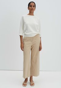 Donna in piedi, con indosso una maglia bianca a maniche tre quarti, pantaloni beige a gamba larga e sandali infradito beige su uno sfondo semplice.