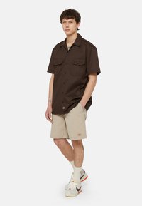 Brown short-sleeve button-up shirt with two chest pockets, paired with beige shorts. Model wears white sneakers and light-colored socks.