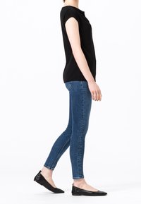 Person som står på sidan i en svart t-shirt med kort ärm, blå smala jeans och svarta platta skor mot en vit bakgrund.