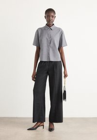 Gray short-sleeve button-up shirt paired with loose black trousers. Model holds a black handbag with chain detail and wears black heels.