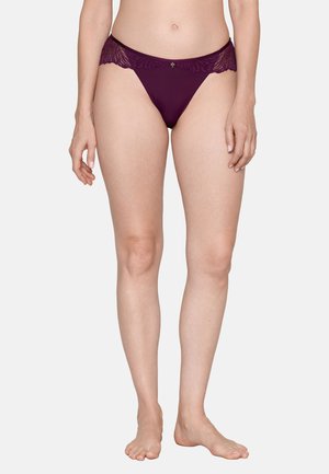 Vrouw die een donkerpaarse bikini met kanten afwerking draagt, staat blootsvoets tegen een eenvoudige lichte achtergrond.