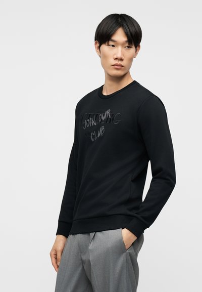 Νέος άνδρας με μαύρο φούτερ που γράφει «JOIN OUR CLUB», κοιτάζει αριστερά, με τα χέρια στις τσέπες, φορά ανοιχτό γκρι παντελόνι, μπροστά σε απλό φόντο.