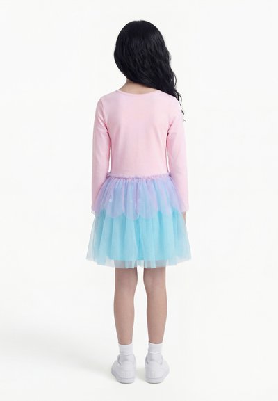 Enfant aux longs cheveux foncés portant un haut rose à manches longues et une jupe en tulle superposée bleue et violette, debout dos à la caméra.