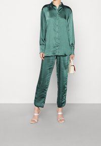Conjunto de pijama de satén en verde esmeralda; camisa de manga larga con cierre de botones, pantalones de pierna ancha; combinado con tacones plateados finos y un bolso pequeño.