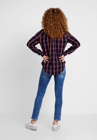 Navy plaid shirt with red accents, button-up design, long sleeves, and curved hem, paired with blue skinny jeans and white sneakers.