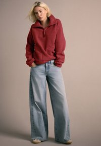 Tânără purtând un pulover roșu din fleece cu fermoar și blugi largi, albaștri deschis, stând cu mâinile în buzunare pe un fundal neutru.
