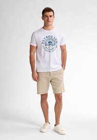 Valkoinen T-paita, jossa on sininen aurinkokuvio ja teksti; yhdistetty beigen värisiin shortseihin ja vaaleisiin tennareihin. Malli seisoo rennosti käsivarret alas.