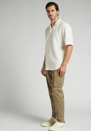 Kortærmet, cremefarvet skjorte med krave og lodrette striber; parret med tætsiddende, khaki cargo-bukser og lyse sneakers.