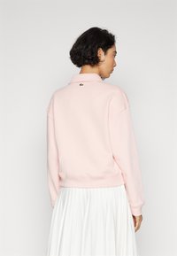Sudadera rosa con cuello y hombros caídos, con un pequeño logo verde en la parte trasera. El material parece suave y cuenta con puños acanalados.
