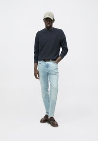 Marineblå striktrøje, lyseblå straight-leg jeans, brune snøresko og en beige kasket. Modellen poser foran en ensfarvet hvid baggrund.