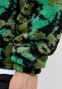 Veste en polaire verte, noire et olive avec une surface texturée et duveteuse et un motif camouflage. Dispose de manches roulées mettant en valeur le tissu.