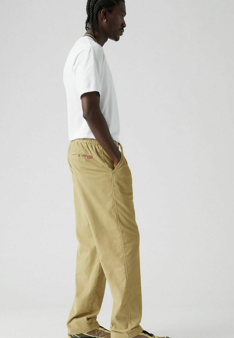 Pantalones jogger de algodón beige con una cinturilla elástica y bolsillos laterales, con un pequeño logo rojo y diseño de pierna recta. Combinados con una camiseta blanca.