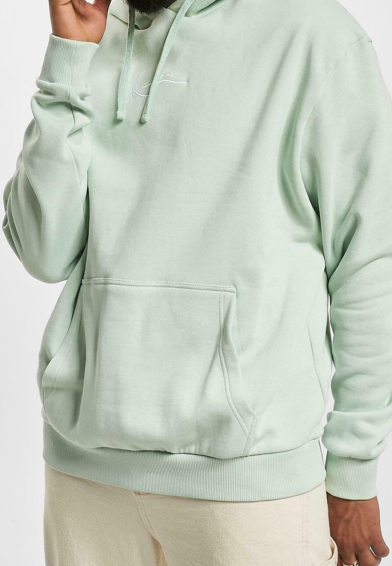 Homme portant un sweat à capuche vert clair avec poche avant et cordons, associé à un pantalon beige clair, main près du visage.