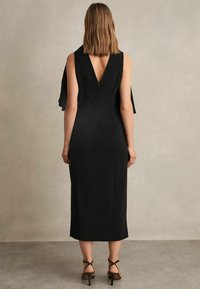 Schwarzes ärmelloses Kleid mit V-Ausschnitt, figurbetontem Schnitt und bodenlangem Saum. Kombiniert mit geflochtenen High Heels.