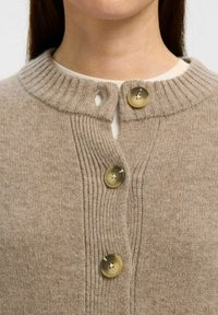 Gilet en tricot beige avec col côtelé, comportant deux gros boutons ronds et une texture douce. Gros plan sur l'encolure et les boutons.