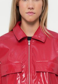 Giacca in ecopelle rossa e lucida con chiusura a zip, colletto a punta e due tasche sul petto con bottoni a scatto.