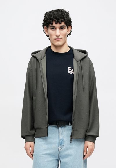 Γκρι φούτερ με φερμουάρ και υφή, με κουκούλα και μπροστινές τσέπες, φορεμένο πάνω από μπλε πουλόβερ με λογότυπο "EA", συνδυασμένο με ανοιχτόχρωμο τζιν.