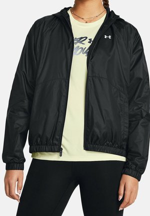 Person trägt eine schwarze Kapuzenjacke mit Reißverschluss über einem hellgelben Under Armour Shirt und schwarzen Leggings.